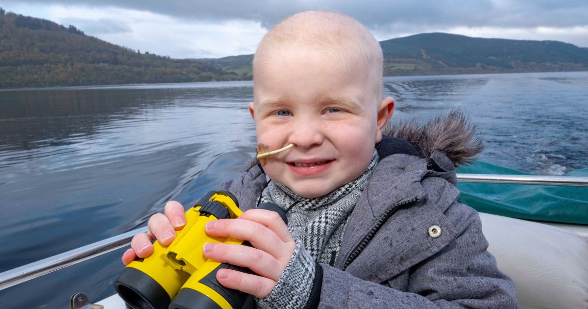 Young boy smiling to camera holding a pair of binoculars. There is a loch and green hills behind him