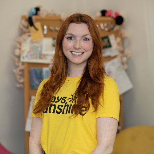 Clodagh is wearing a yellow Rays of Sunshine t-shirt. She is standing. Clodagh has ginger hair