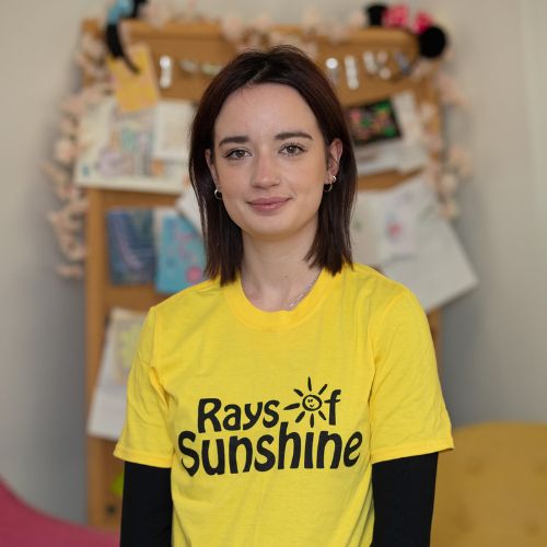 Beth is standing and is wearing a Rays of Sunshine t-shirt. She has black hair.