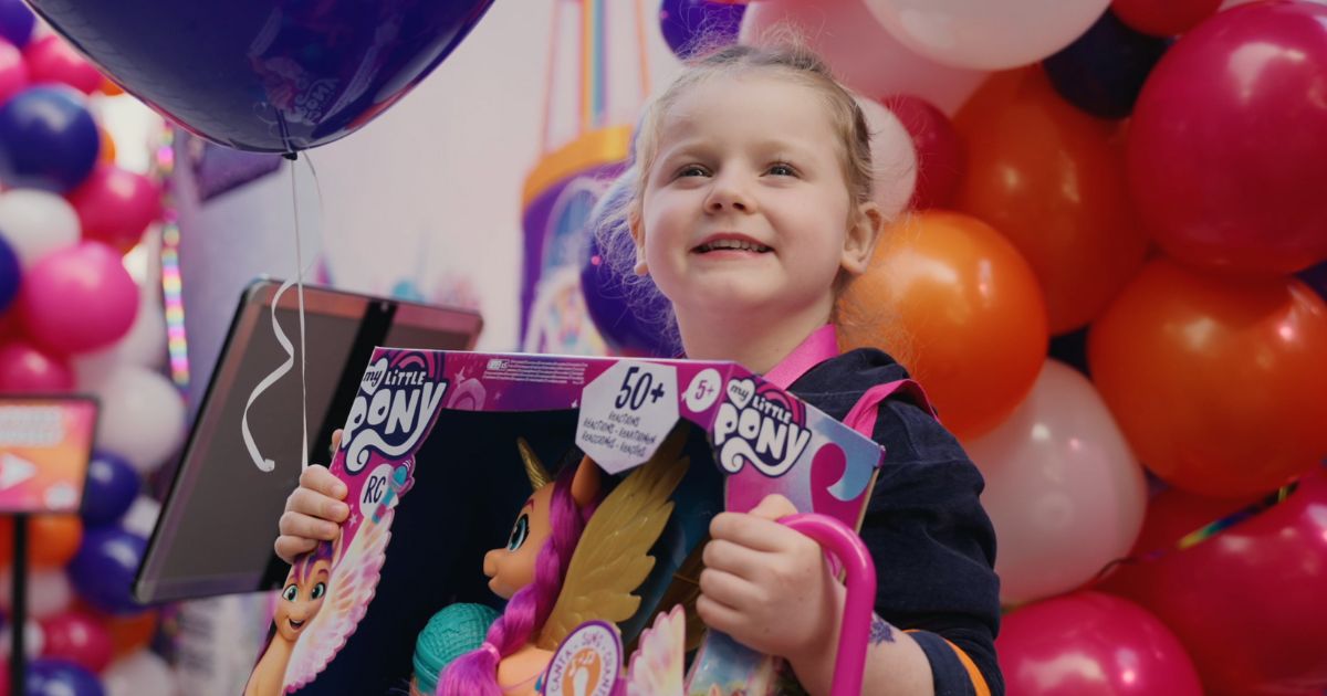 A girl smiling holding a box of My Little Pony toys. There are colourful balloons in the background