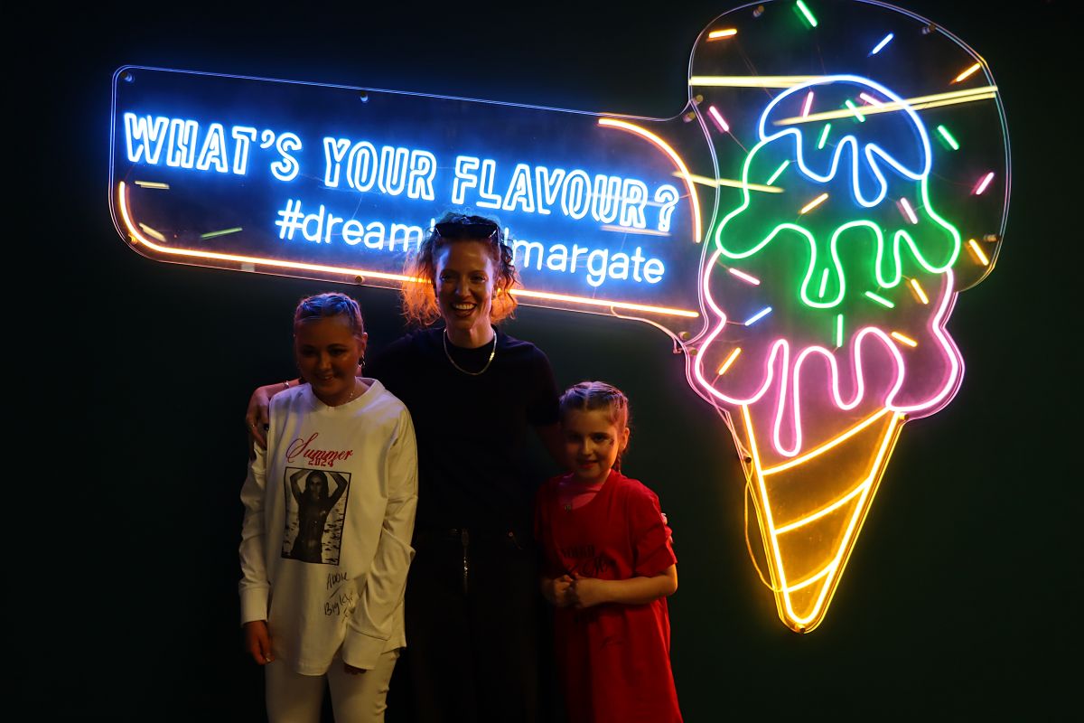 Three people smiling for a pic in front of a bright neon colour sign. There are two younger girls/teens and an older woman.