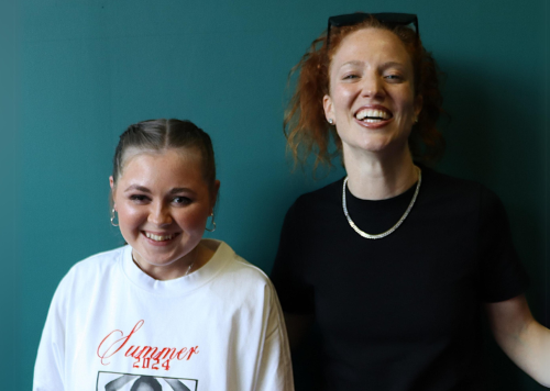 Two people smile while hugging each other and posing for a pic in front of a green wall. One is a young teen and the other a woman