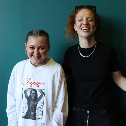 Two people smile while hugging each other and posing for a pic in front of a green wall. One is a young teen and the other a woman