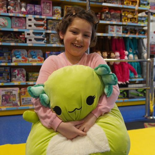 Little girl smiles for the camera while cuddling a big green Squishmallow. She is stood in a brightly lit toy shop