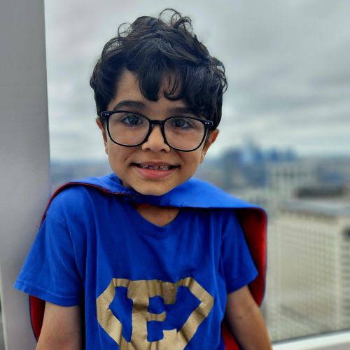 Little boy smiles for the camera. He has thick glasses and brown hair. He is wearing a blue t-shirt with a superhero emblem with an E on it.