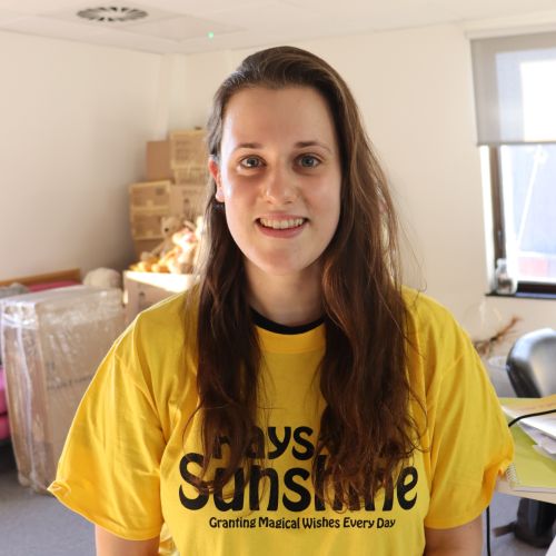 Woman with long dark blonde hair smiles while stood in an office. She wears a yelow t-shirt