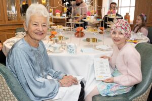 A little girl and an older woman smile for the camera. They have turned away from a table which is laid out with tea supplies.