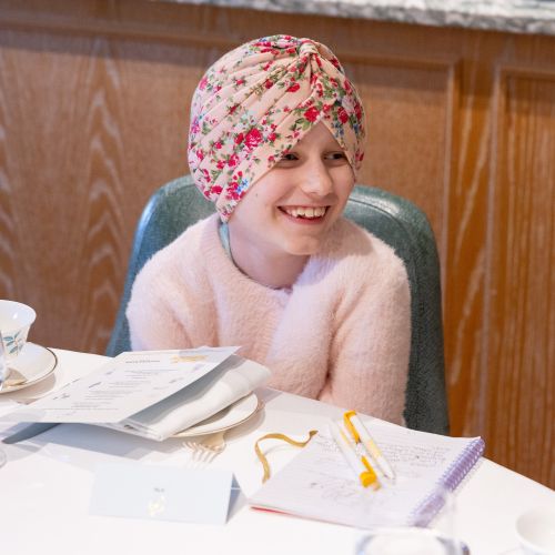 Little girl wearing flowery head scarf smiles at something off camera. She is sat at a table wih a notebook open