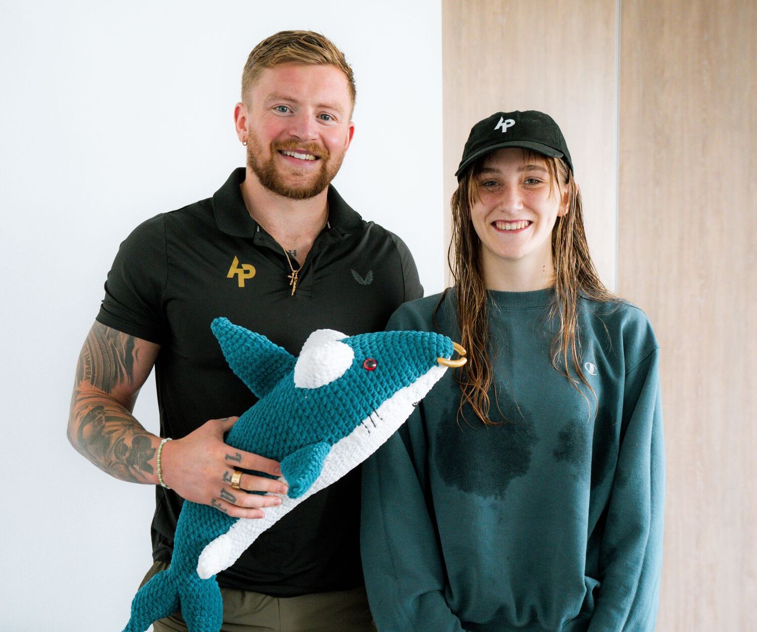 Two people smiling for a picture. One is an adult man, who wears a dark coloured t-shirt and shorts. He holds an oversize crochet stuffed shark. The other person is a teen girl, who wears a green sweatshirt and joggers. She also has wet hair and a baseball cap on.