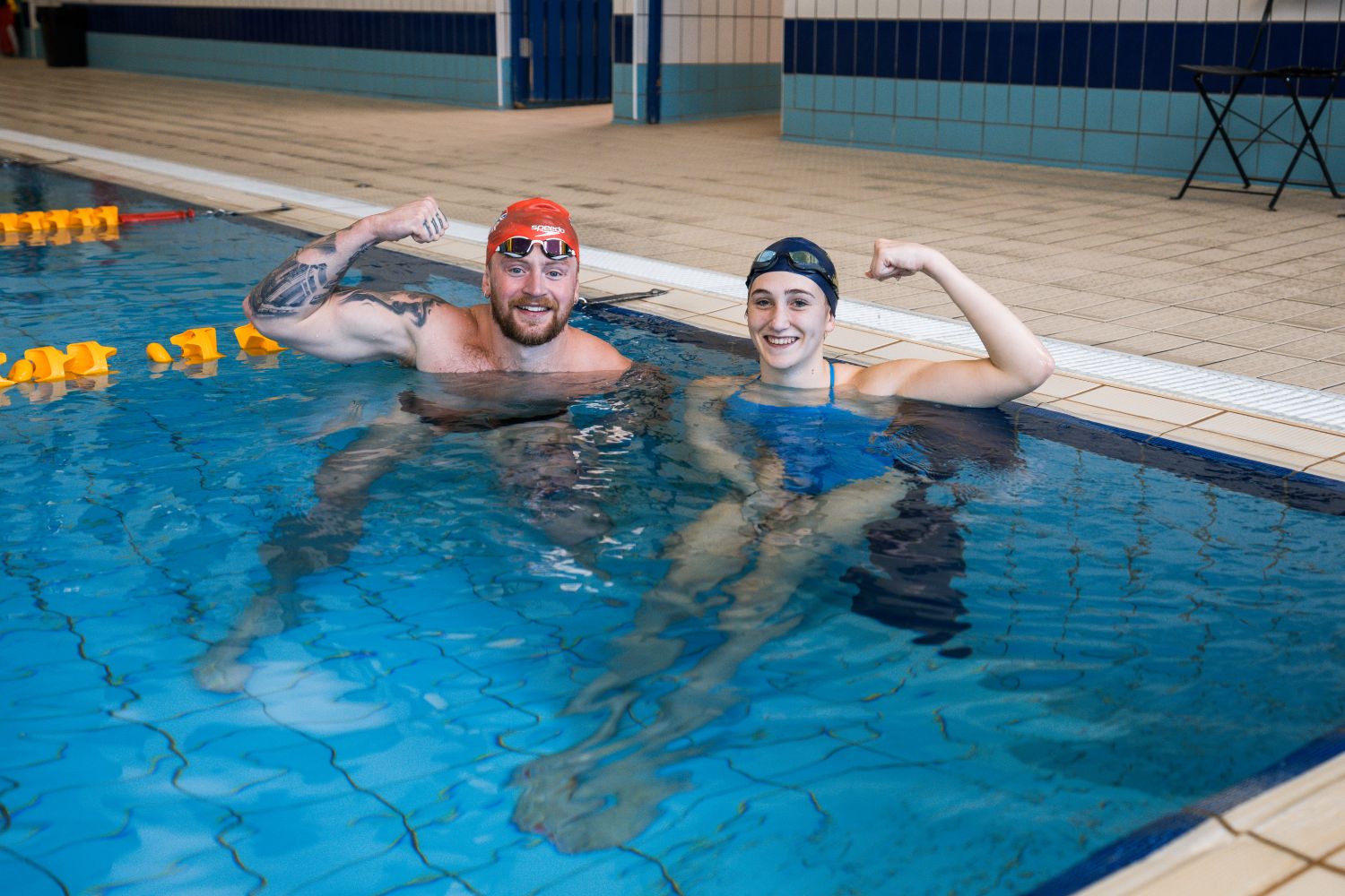 Two people stand in an indoor swimming pool. They both wear swimming caps, and are posing with an arm held in a "strong" pose