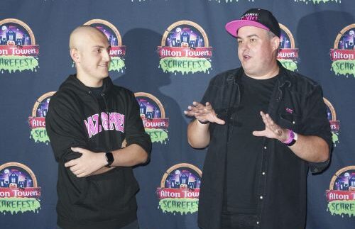 Two people stand while talking to each other. They are stood in front of a press board featuring the Alton Towers logo. They both wear dark clothes, with the older of the two also wearing a baseball cap