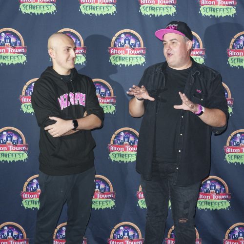 Two people stand while talking to each other. They are stood in front of a press board featuring the Alton Towers logo. They both wear dark clothes, with the older of the two also wearing a baseball cap