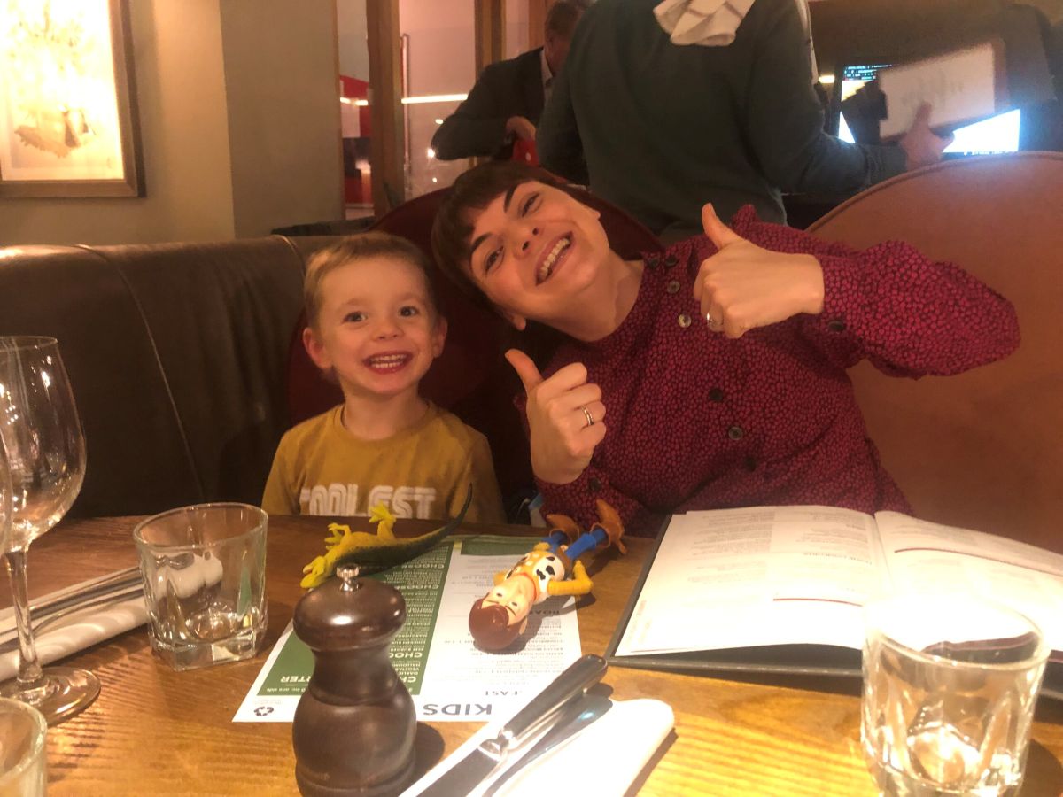 Little boy and his mum sit at the dinner table. They are in a restaurant, and both smile at the camera. The mum holds up two thumbs-up and tilts her head to the little boy.