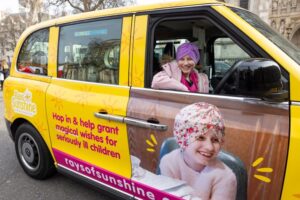 Little girl sits in the driver's seat of a bright yellow taxi while leaning out and smiling at the camera. 