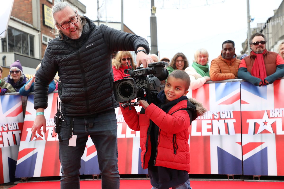 Little boy holding a video camera in front of a group of people lined up for a red carpet