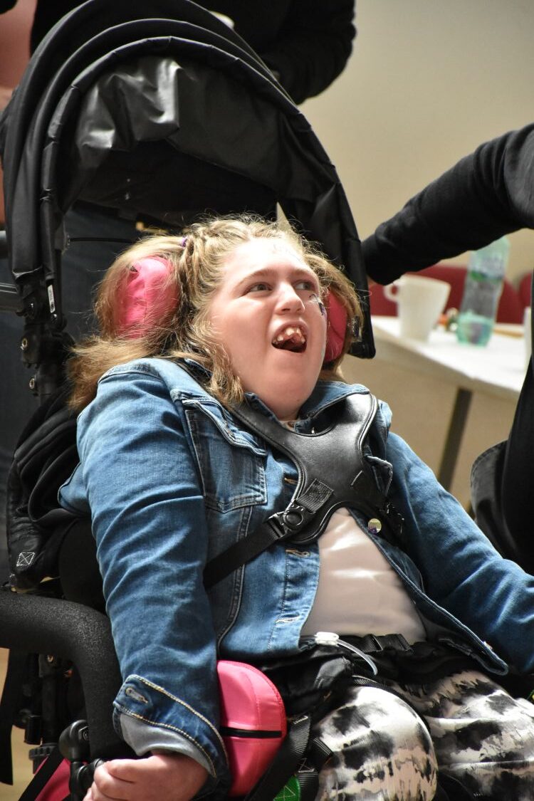 Girl in a wheelchair smiles at people out of frame.