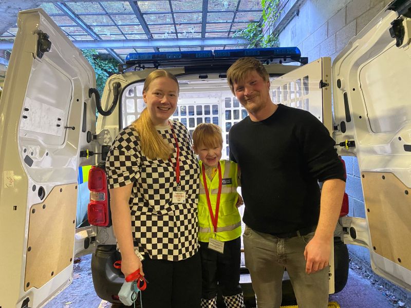Parents and little boy smile while stood at the back of a police van
