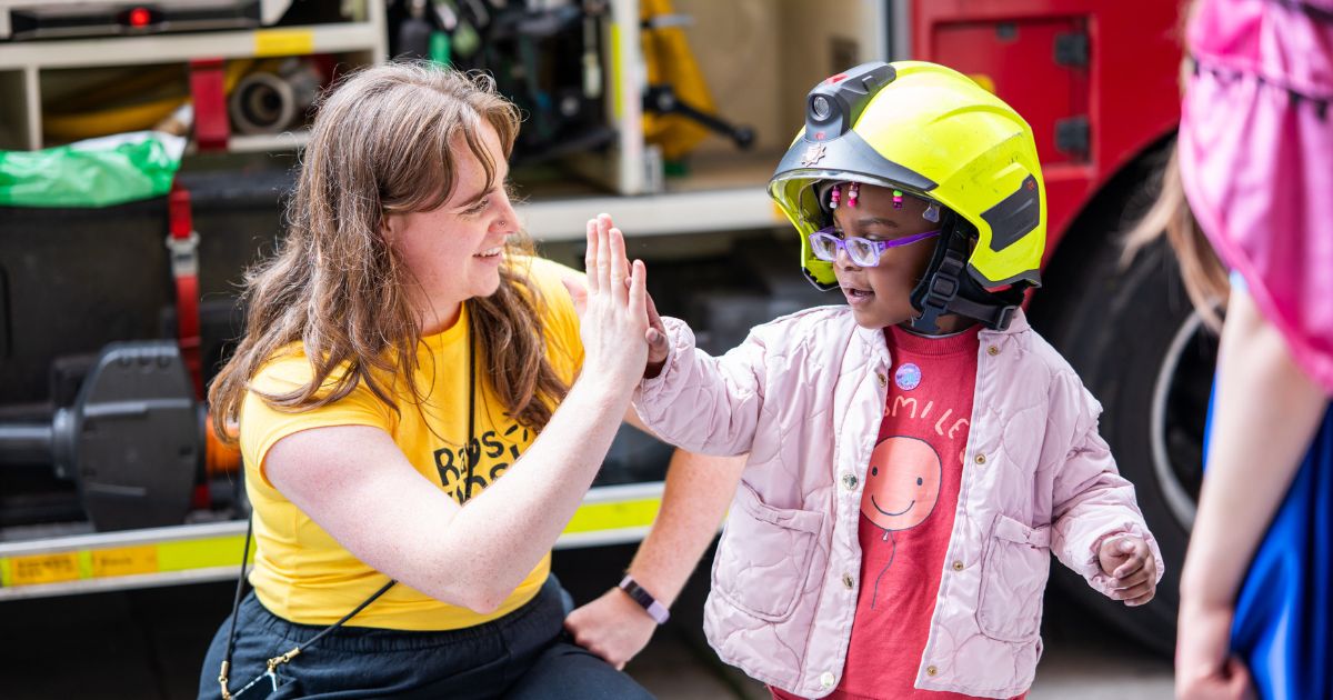 Woman in Rays of Sunshine t-shirt high fives little girl wearing firefighter helmet