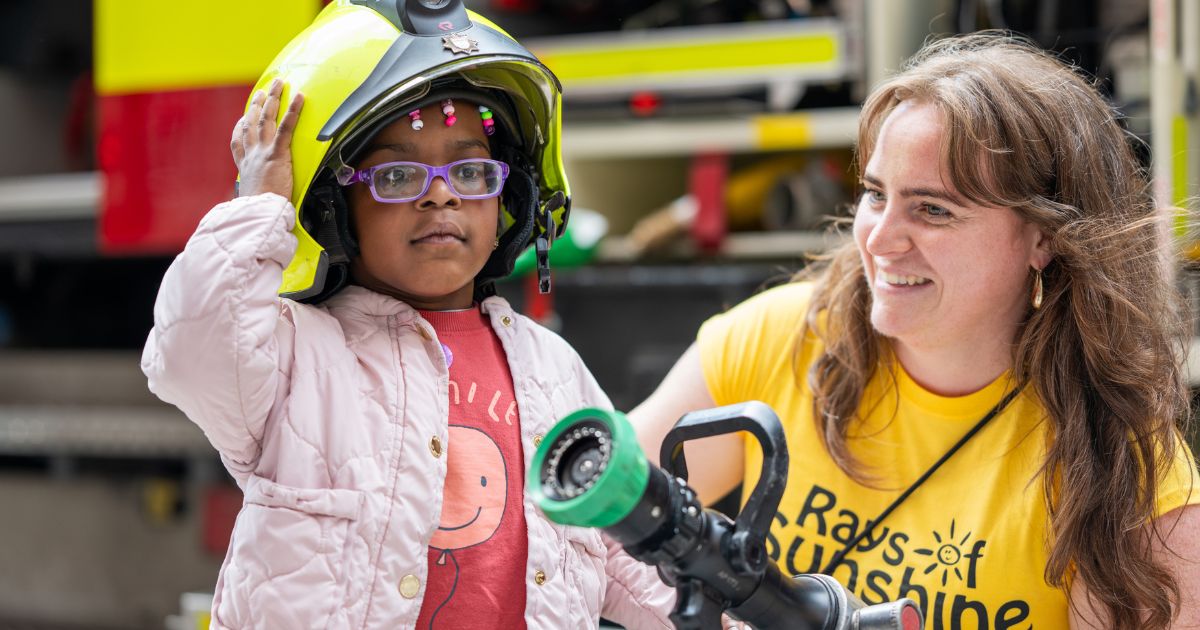 Little girl with firefighter hat on with woman in rays of sunshine t-shirt holding a fire hose