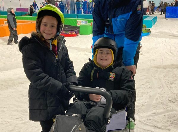 Brother and sister smiling for the camera while on an indoor ski slope. The boy sits in an adaptive ski sled, and the girl stands beside her.