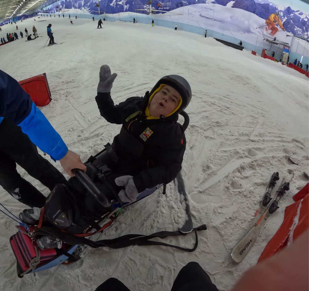Boy posing for a photo while sat in an adaptive skiing sled while at an indoor ski slope