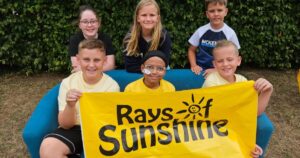 Six smiling children pose with a yellow "Rays of Sunshine" banner. One child, marked with yellow lines, wears a nasal cannula.