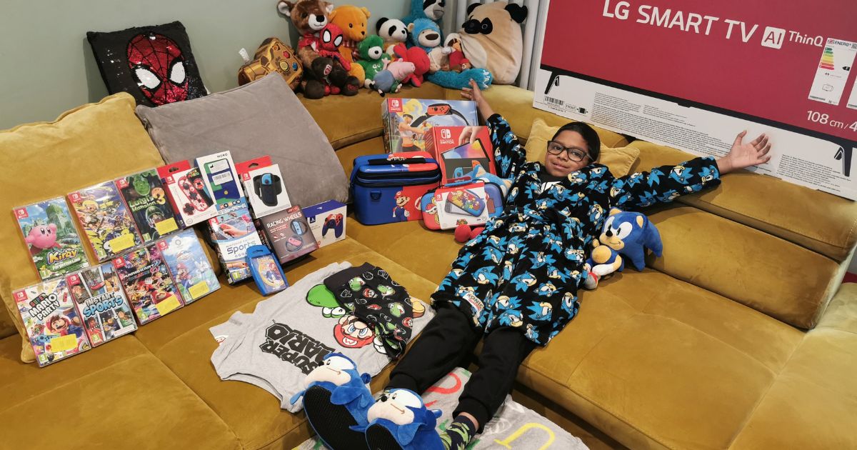 Smiling child on a yellow couch surrounded by toys, games, and a large LG Smart TV. Wearing a Sonic robe, slippers, and socks.