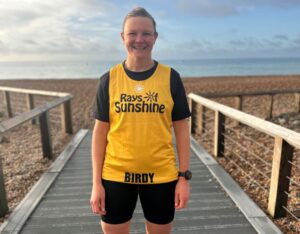 Woman stands by the sea, while wearing a yellow running vest which reads: Rays of Sunshine