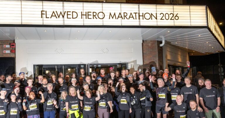 A group of runners smiling and cheering in front of a sign that says Flawed Hero Marathon 2026