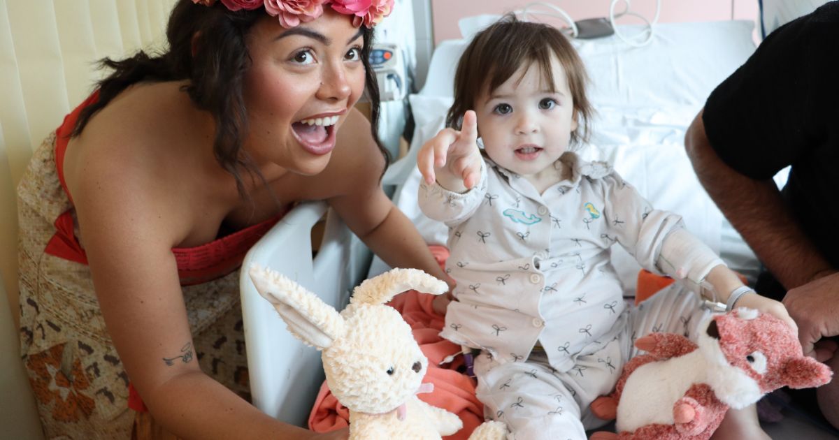 Young girl in hospital bed pointing to the camera with woman dressed as Moana doing a shocked face